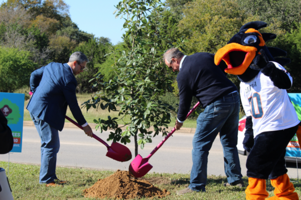 two people planting a new tree; Rowdy stands nearby in his Rowdy jersey