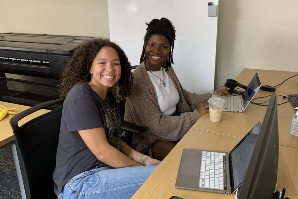 two students sitting together at a desk smiing and lolking up form a computer screen